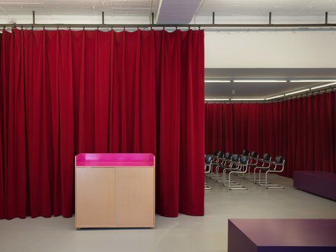  "Ein Innenraum, der durch rote Samtvorhänge zweigeteilt ist. Im Vordergrund steht ein helles Holzpult mit einer leuchtend pinkfarbenen Ablagefläche. Links hängen blickdichte rote Vorhänge. Rechts öffnet sich der Raum, wo hinter einem Teil der roten Vorhänge Reihen von schwarzen Stühlen für eine Veranstaltung aufgestellt sind."