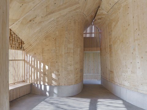 "Innenansicht einer zylindrischen Holzstruktur mit gebogenem Gewölbe aus verleimtem Sperrholz. Gekrümmte Holzrahmen verlaufen von oben nach unten. Vertikale Lamellen erzeugen Licht- und Schattenmuster. Helles Sperrholz mit natürlichen Astlöchern. Moderne architektonische Details der Holzkonstruktion."