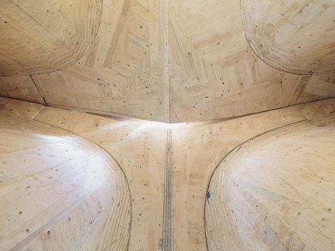 "Blick von unten auf die gewölbte Holzdecke einer zylindrischen Struktur. Zwei große gekrümmte Holzrahmen aus verleimtem Sperrholz treffen sich in der Mitte. Das helle Sperrholz zeigt natürliche Astlöcher und Unebenheiten. Schmale vertikale Lamellen trennen die beiden Gewölbehälften. Sonnenlicht fällt durch kleine Öffnungen und erzeugt feine Schattenstreifen auf der Holzoberfläche."