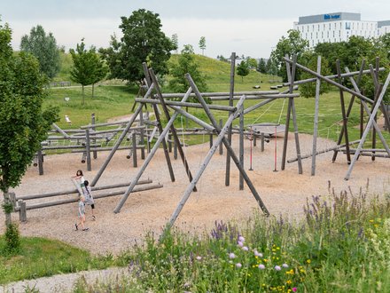 Ein Spielplatz mit Klettergerüsten aus Holz, auf dem zwei Kinder spielen. Im Hintergrund sind Bäume und ein Gebäude zu sehen. Der Boden des Spielplatzes ist mit Kies bedeckt, und bunte Blumen wachsen am Rand.