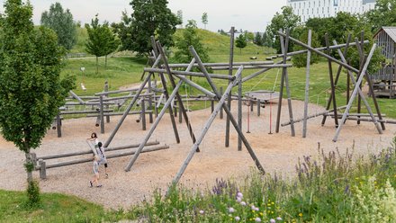 Ein Spielplatz mit Klettergerüsten aus Holz, auf dem zwei Kinder spielen. Im Hintergrund sind Bäume und ein Gebäude zu sehen. Der Boden des Spielplatzes ist mit Kies bedeckt, und bunte Blumen wachsen am Rand.