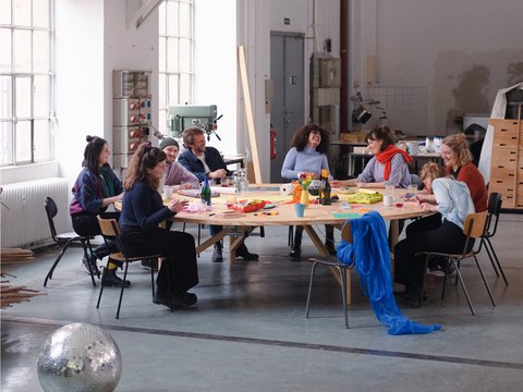Neun diverse Personen sitzen um einen großen runden Holztisch in einer hellen Industriewerkstatt. Sie arbeiten an kreativen Projekten mit Materialien und Kunsthandwerk. Ein silberner Discokugel liegt auf dem Betonboden im Vordergrund. Große Fenster spenden natürliches Licht, im Hintergrund sind Werkzeuge und Maschinen zu sehen."