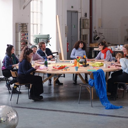 Neun diverse Personen sitzen um einen großen runden Holztisch in einer hellen Industriewerkstatt. Sie arbeiten an kreativen Projekten mit Materialien und Kunsthandwerk. Ein silberner Discokugel liegt auf dem Betonboden im Vordergrund. Große Fenster spenden natürliches Licht, im Hintergrund sind Werkzeuge und Maschinen zu sehen."