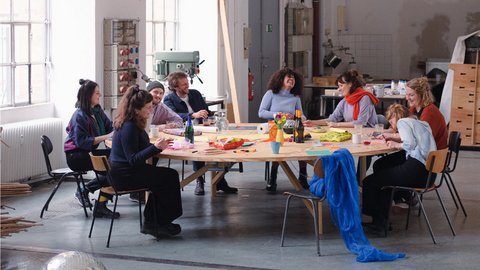 Neun diverse Personen sitzen um einen großen runden Holztisch in einer hellen Industriewerkstatt. Sie arbeiten an kreativen Projekten mit Materialien und Kunsthandwerk. Ein silberner Discokugel liegt auf dem Betonboden im Vordergrund. Große Fenster spenden natürliches Licht, im Hintergrund sind Werkzeuge und Maschinen zu sehen."