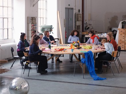 Neun diverse Personen sitzen um einen großen runden Holztisch in einer hellen Industriewerkstatt. Sie arbeiten an kreativen Projekten mit Materialien und Kunsthandwerk. Ein silberner Discokugel liegt auf dem Betonboden im Vordergrund. Große Fenster spenden natürliches Licht, im Hintergrund sind Werkzeuge und Maschinen zu sehen."