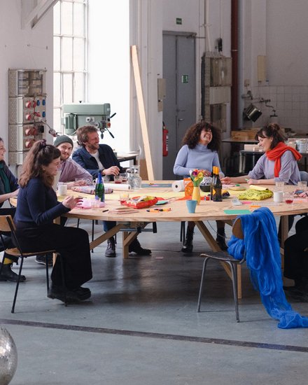 Neun diverse Personen sitzen um einen großen runden Holztisch in einer hellen Industriewerkstatt. Sie arbeiten an kreativen Projekten mit Materialien und Kunsthandwerk. Ein silberner Discokugel liegt auf dem Betonboden im Vordergrund. Große Fenster spenden natürliches Licht, im Hintergrund sind Werkzeuge und Maschinen zu sehen."