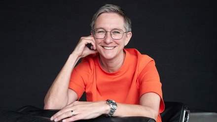 "Porträt eines Mannes mit Brille und grauem Haar, der ein leuchtendes rotes T-Shirt trägt. Er sitzt vor dunklem Hintergrund, lächelt in die Kamera und stützt seinen Kopf entspannt mit der linken Hand ab. Er trägt eine silberne Uhr und einen Ring."