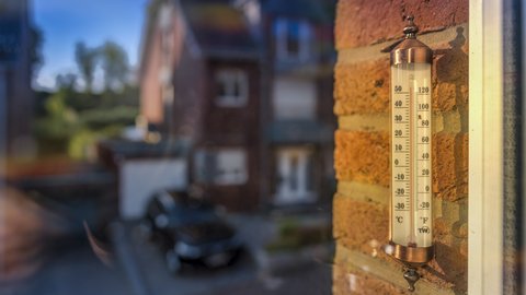 "Nahaufnahme eines Thermometers mit kupfernem Gehäuse an einer rötlichen Backsteinmauer montiert. Das Thermometer zeigt etwa 28°C an. Im unscharfen Hintergrund sind historische Gebäude unter blauem Himmel zu sehen."