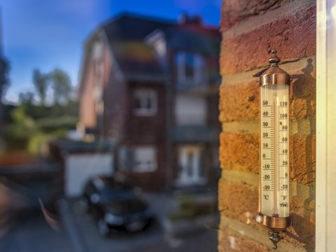 "Nahaufnahme eines Thermometers mit kupfernem Gehäuse an einer rötlichen Backsteinmauer montiert. Das Thermometer zeigt etwa 28°C an. Im unscharfen Hintergrund sind historische Gebäude unter blauem Himmel zu sehen."