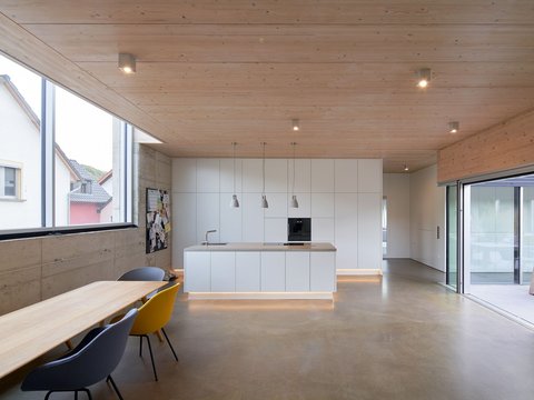 Moderner Küchen- und Essbereich mit freistehender, bodentief beleuchteter Kochinsel, Holzdecke, Sichtbetonwänden und großen Fensterfronten.