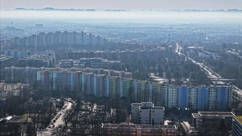 "Weitläufiger Blick von oben über ein städtisches Wohngebiet. Langgestreckte, teils pastellfarbene Hochhauskomplexe ziehen sich durch das Bild. Im diesigen Hintergrund ist eine Bergkette am Horizont zu erkennen."