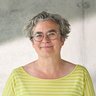 "Porträt einer Frau mit grauem, lockigem Haar und Brille. Sie trägt ein gelb-weiß gestreiftes Langarmshirt und lächelt in die Kamera. Im Hintergrund eine helle Betonwand mit Fenster und natürlichem Licht."