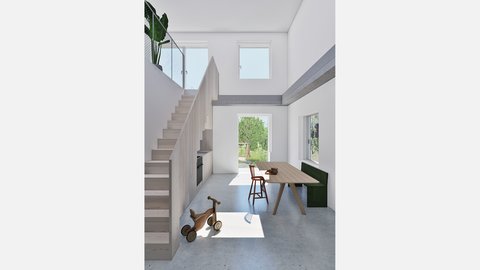 Wohnraum mit Treppe und Gartentür Alt: Doppelgeschossiger Raum mit Holztreppe und integrierter Küchenzeile; großer Esstisch; Gartentür im Gegenlicht; Holz‑Dreirad am Boden.