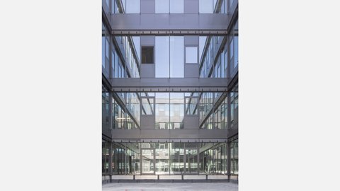 Innenhof mit Glasfassaden  Ein mehrgeschossiger Innenhof, umgeben von Glas- und Metallfassaden. Fenster und Brüstungen spiegeln sich gegenseitig. Der Boden ist leer, der Raum wirkt ruhig und geschlossen. Das Bild zeigt einen gemeinschaftlichen Innenraum, der Licht ins Gebäude bringt und visuelle Verbindungen zwischen den Ebenen schafft.