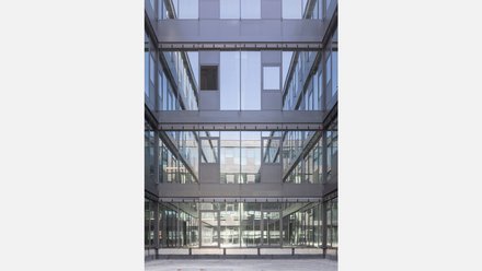 Innenhof mit Glasfassaden  Ein mehrgeschossiger Innenhof, umgeben von Glas- und Metallfassaden. Fenster und Brüstungen spiegeln sich gegenseitig. Der Boden ist leer, der Raum wirkt ruhig und geschlossen. Das Bild zeigt einen gemeinschaftlichen Innenraum, der Licht ins Gebäude bringt und visuelle Verbindungen zwischen den Ebenen schafft.