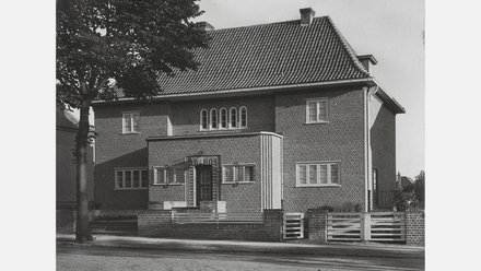 Schwarzweiß-Fotografie eines zweistöckigen Backsteinhauses mit Satteldach und Ziegeln. Das Haus hat symmetrisch angeordnete Fenster, eine weiße Veranda mit Stufen zum Eingang und einen Baum links davon. Das Gebäude wirkt wie ein klassisches deutsches Wohnhaus aus der Nachkriegszeit.