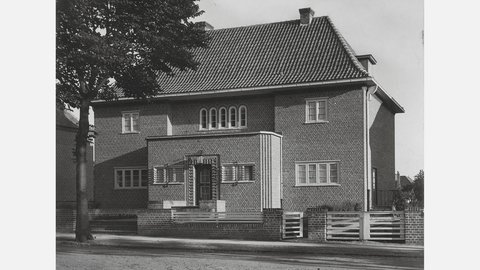 Schwarzweiß-Fotografie eines zweistöckigen Backsteinhauses mit Satteldach und Ziegeln. Das Haus hat symmetrisch angeordnete Fenster, eine weiße Veranda mit Stufen zum Eingang und einen Baum links davon. Das Gebäude wirkt wie ein klassisches deutsches Wohnhaus aus der Nachkriegszeit.