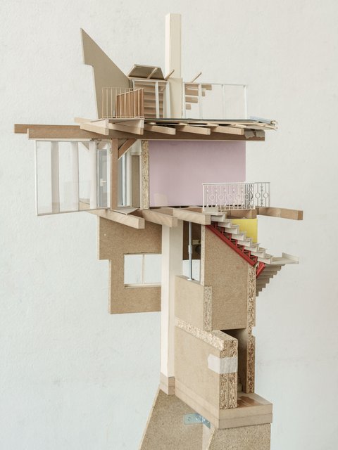 Diese Seitenansicht des gleichen Modells bietet tiefere Einblicke in die Innenraumgestaltung und Materialität. Sichtbar sind verschiedene Wandabschnitte aus rosa gestrichenem Material und Holz, eine Treppe aus Sperrholz mit roter Wange, eine Balkonbrüstung aus filigranem Metall und große Glasfassadenelemente. Der offene, fragmentierte Charakter des Modells wird durch die freiliegenden Holzkonstruktionen betont. Der hohe Sockel aus Spanplatten ist hier ebenfalls zu sehen. Der Hintergrund ist wiederum die weiße Wand mit dem gefliesten Bereich.