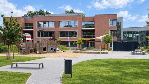 Moderner Backsteinbau einer Grundschule mit Spielplatz, Rasenfläche und gepflastertem Weg im Vordergrund