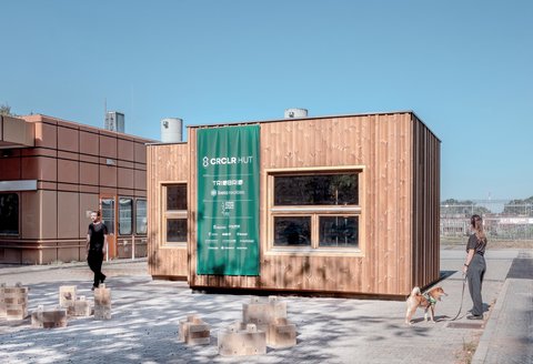 Holzmodul mit großen Fenstern und grünem Banner, Menschen und Holzbausteine davor.
