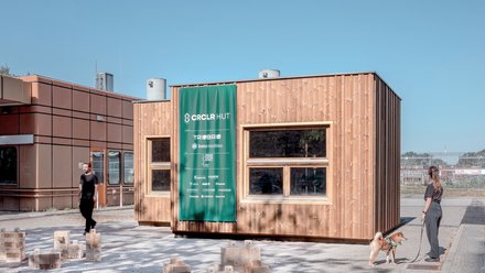 Holzmodul mit großen Fenstern und grünem Banner, Menschen und Holzbausteine davor.