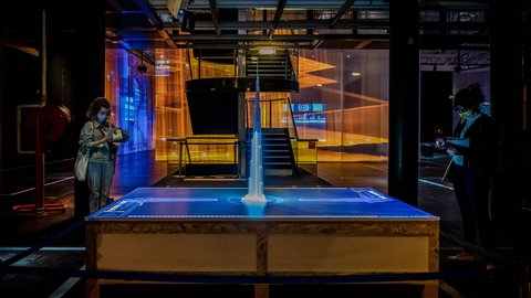 "Dunkler Ausstellungsraum mit interaktiver Installation. Auf einem Tisch steht ein beleuchtetes Architekturmodell eines hohen Gebäudes. Die Oberfläche des Tisches zeigt digitale Projektionen in Blau. Zwei Besucher stehen seitlich und fotografieren oder betrachten das Modell. Im Hintergrund verlaufen orangefarbene Lichtprojektionen entlang halbtransparenter Wände und einer Treppe."