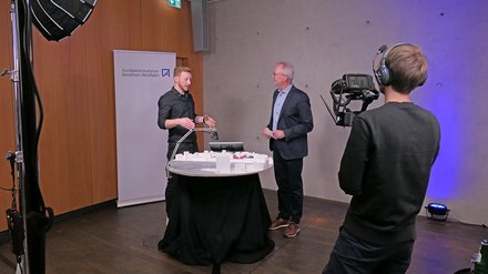 Drehsituation: Zwei Personen erklären ein Architekturmodell am runden Tisch; vorn filmt eine Kameraperson, Softbox‑Licht und Banner „Architektenkammer Nordrhein‑Westfalen“ sichtbar.