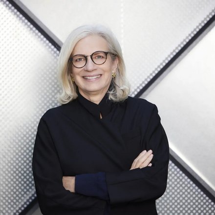 Porträt einer Frau mit heller Brille und verschränkten Armen vor geometrisch gemustertem Hintergrund.