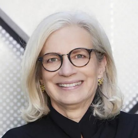 Porträt einer Frau mit heller Brille und verschränkten Armen vor geometrisch gemustertem Hintergrund.