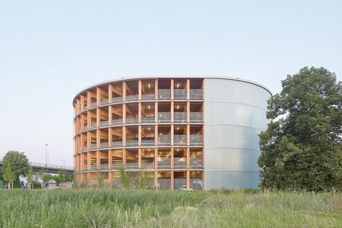 Rundes Parkhaus aus Holz mit offener Fassadenstruktur und transluzenter Hülle, umgeben von Grünfläche.