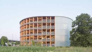 Rundes Parkhaus aus Holz mit offener Fassadenstruktur und transluzenter Hülle, umgeben von Grünfläche.