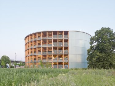 Rundes Parkhaus aus Holz mit offener Fassadenstruktur und transluzenter Hülle, umgeben von Grünfläche.