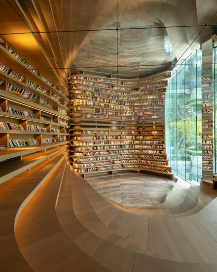 Moderne Bibliothek mit geschwungenen Holzregalen und langen Reihen voller Bücher. Rechts öffnen hohe Glaswände den Blick in einen grünen Garten. Warmes Licht betont die fließende Architektur und die ruhige, offene Raumwirkung.