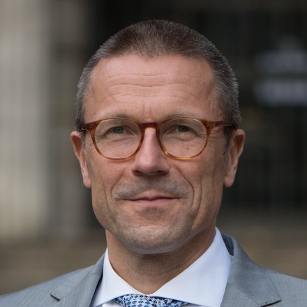 Porträt eines Mannes mit Brille im grauen Anzug und gemusterter Krawatte vor unscharfem Hintergrund.