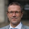 Porträt eines Mannes mit Brille im grauen Anzug und gemusterter Krawatte vor unscharfem Hintergrund.