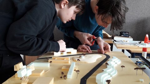 "Zwei Jungs arbeiten konzentriert an einem großen städtebaulichen Modell auf einem Holztisch. Der linke Junge im schwarzen Pullover schaut genau zu, während der rechte Junge im blauen Pullover mit einer kleinen Schere ein schwarzes Band zuschneidet, das eine Straße darstellen könnte. Das Modell zeigt quaderförmige Gebäude aus gelbem Schaumstoff, geschwungene weiße Elemente und kleine Modellbäume
