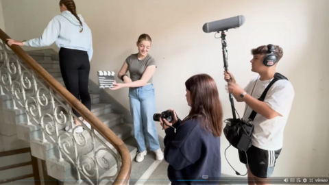 "Ein Stillleben aus einem Video, das Dreharbeiten in einem Treppenhaus zeigt. Ein Mädchen im grauen T-Shirt hält eine Filmklappe (Clapperboard) in die Kamera, während ein anderes Mädchen mit einer DSLR-Kamera filmt. Rechts hält ein Junge mit Kopfhörern eine Tonangel mit einem Mikrofon hoch. Im Hintergrund geht ein weiteres Mädchen die Treppe hoch."