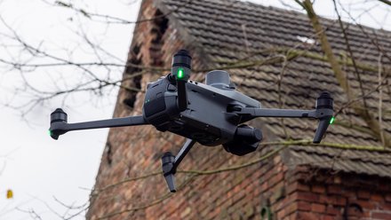 Vierrotorige Drohne mit grünen Positionslichtern fliegt vor einem alten Ziegelgebäude.