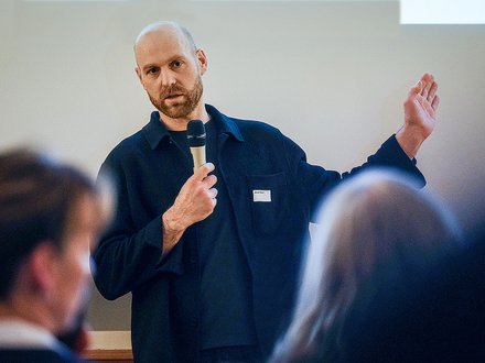 Person mit Glatze und Bart hält ein Mikrofon und spricht vor einem Publikum.