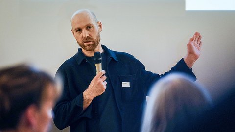 Person mit Glatze und Bart hält ein Mikrofon und spricht vor einem Publikum.