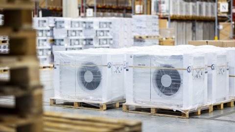 Mehrere verpackte Außeneinheiten von Wärmepumpen stehen auf Paletten in einer Lagerhalle.