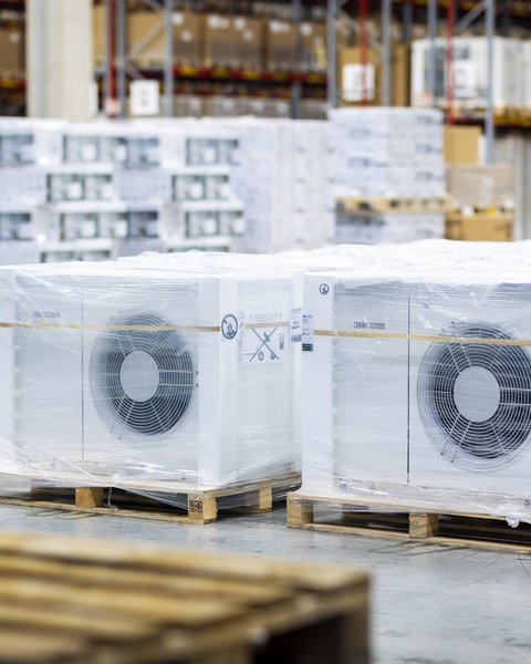 Mehrere verpackte Außeneinheiten von Wärmepumpen stehen auf Paletten in einer Lagerhalle.