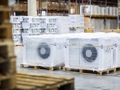 Mehrere verpackte Außeneinheiten von Wärmepumpen stehen auf Paletten in einer Lagerhalle.