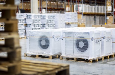 Mehrere verpackte Außeneinheiten von Wärmepumpen stehen auf Paletten in einer Lagerhalle.