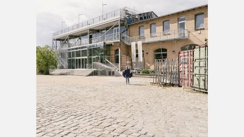 Tagesansicht: gläserner Stahlanbau an historischem Ziegelbau; breite Zugangstreppen, eine Person im Hof, Container rechts.