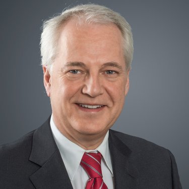 Porträt eines grauhaarigen Mannes im Anzug mit roter Krawatte vor neutralem Hintergrund.