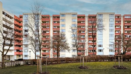 Fassade mit roten Balkonen Alt: Neungeschossiger Plattenbau mit rot abgesetzten Balkonen; davor Rasen und kahle Bäume, blauer Himmel.