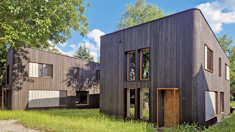 Modernes zweigeschossiges Gebäude mit vertikal verlaufender Holzfassade und großen Fenstern in einer grünen Umgebung