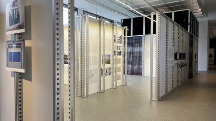 Ein heller Innenraum mit hohen Decken und gleichmäßigem, industriellem Licht. In der Mitte stehen mehrere schmale, senkrechte Metallrahmen mit Lochprofilen, an denen Texttafeln und Bilder befestigt sind. Die Stellwände bilden einen offenen Parcours, durch den Besucher hindurchgehen können. Das Bild zeigt die räumliche Gestaltung einer Ausstellung. Die modulare Konstruktion erinnert an technische Infrastruktur und verweist auf Ordnung, Systematik und Datenstrukturen.