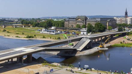 Große Straßenbrücke im Bauzustand über einen Fluss; ein Brückensegment ist abgesenkt, Baumaschinen arbeiten im Wasser und am Ufer, im Hintergrund sind historische Gebäude, Grünflächen und eine Stadtansicht zu sehen.